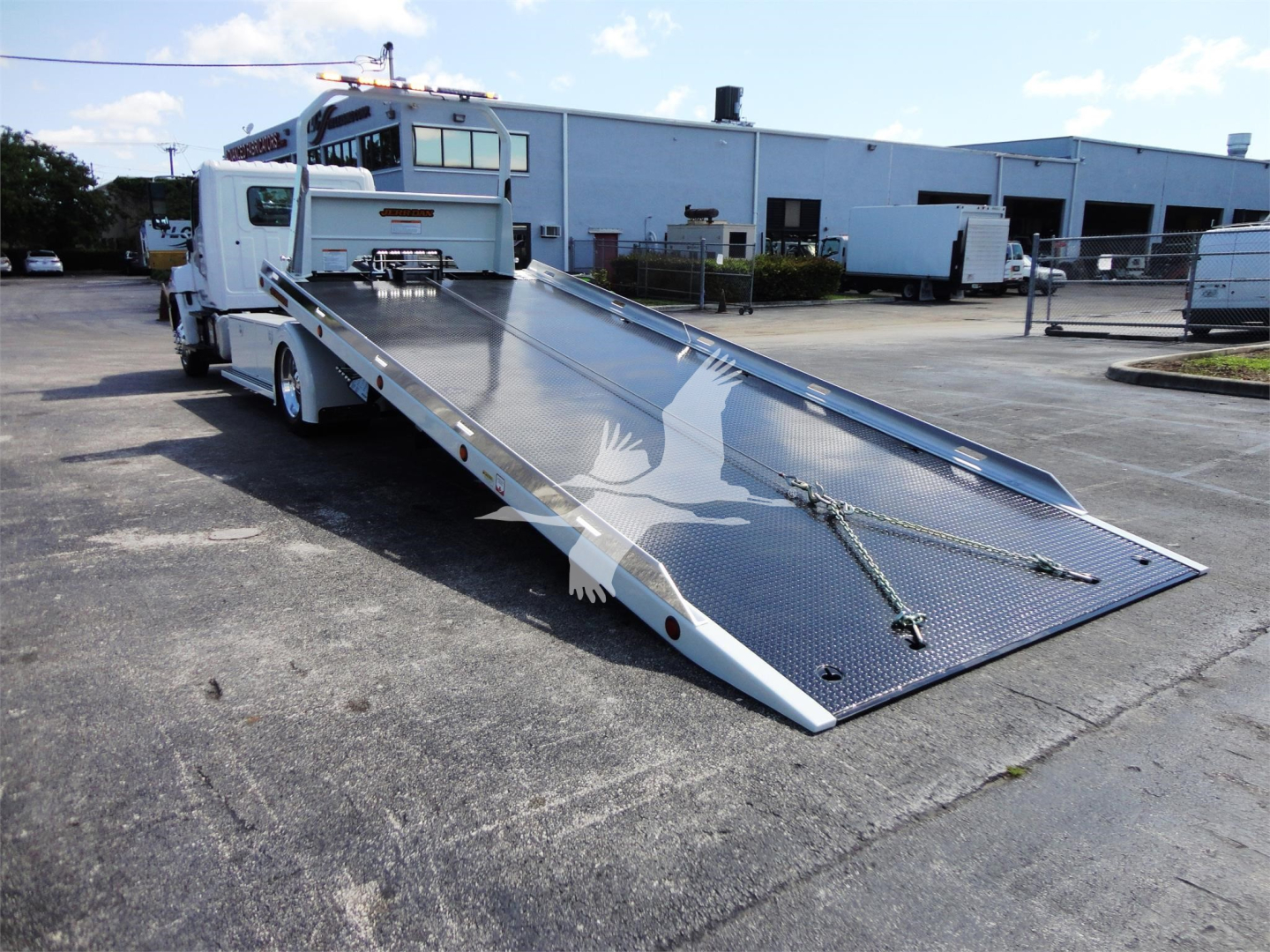 Truck bed of a 2022 Hino 268A rollback tow truck showing a rear tilt flatbed and chrome diamond plate surface in an industrial yard.