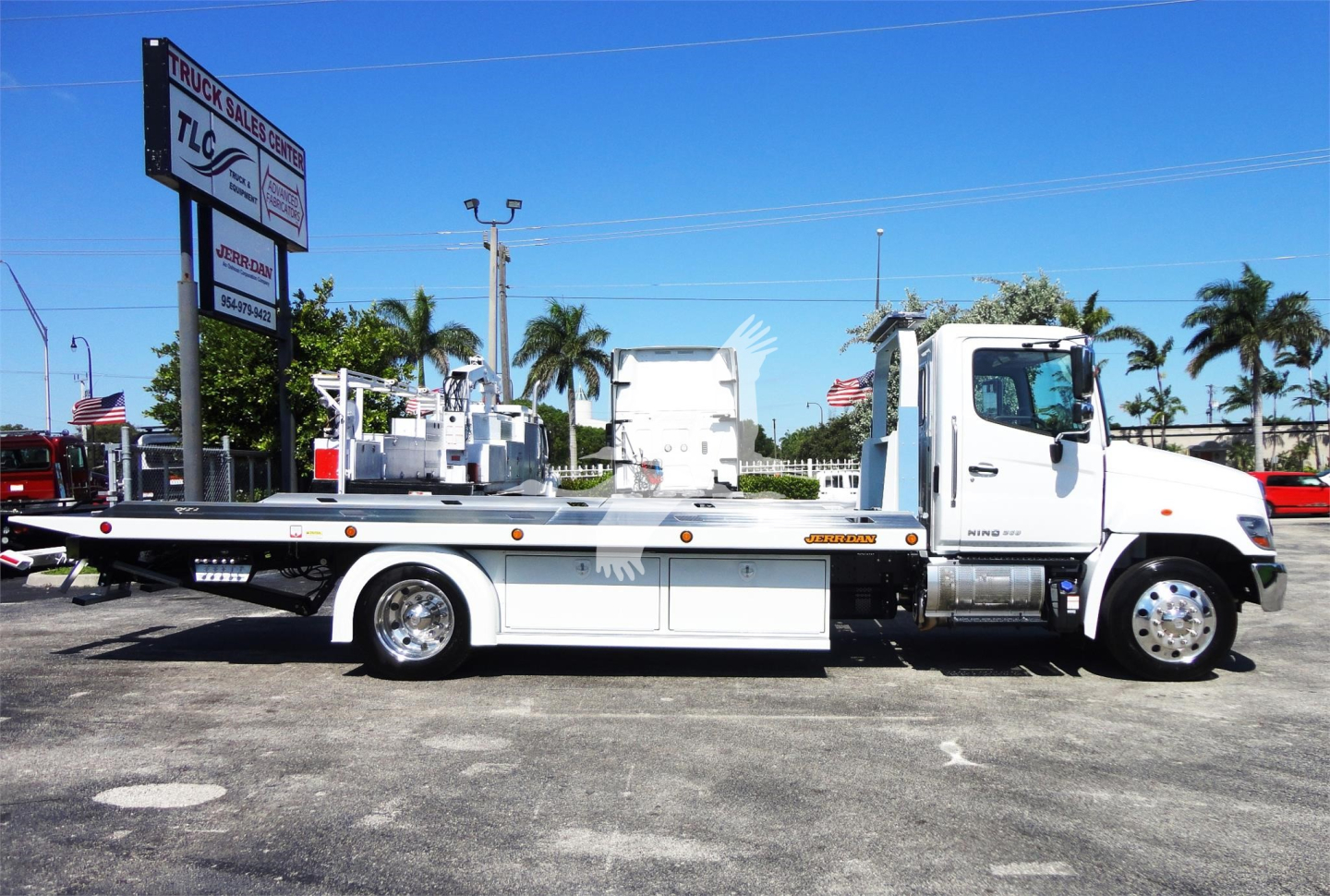 Truck bed rollback tow truck for sale showing silver Hino 268A chassis and rollback bed with side rails and equipment in a dealership lot.