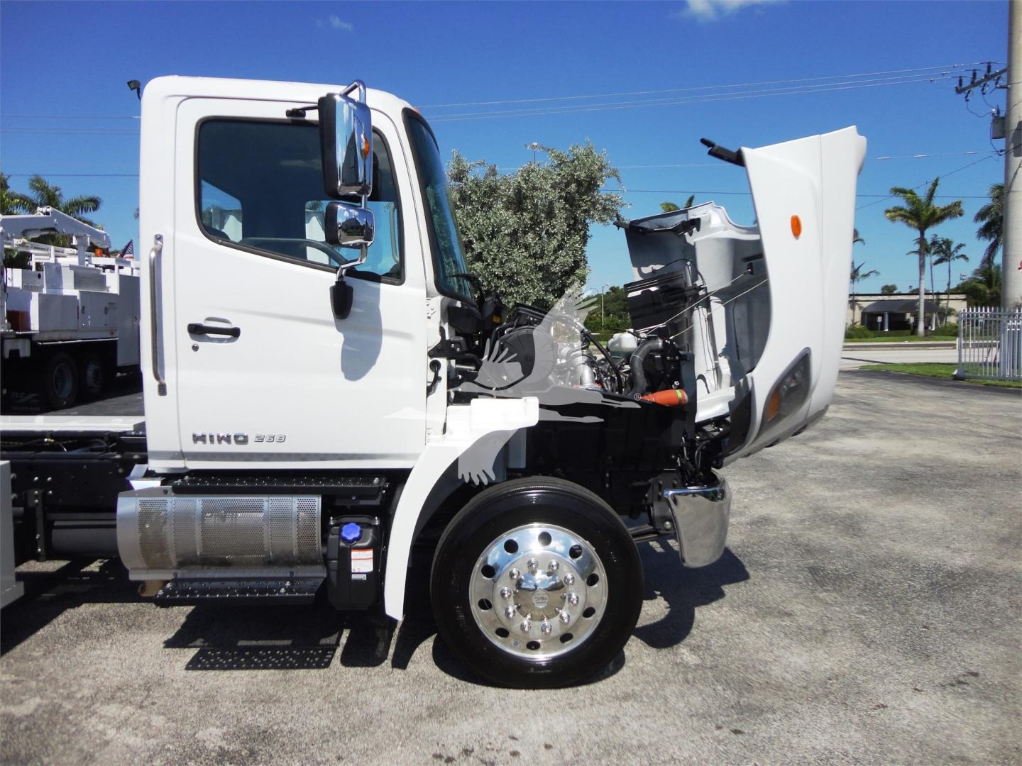 Truck cab of a white 2022 Hino 268A with hood open showing front wheel, chrome hub, and side step. Visible tag area and engine bay.