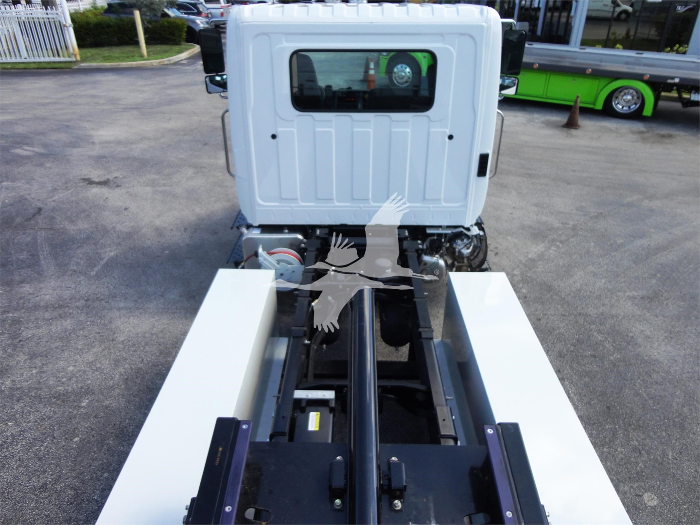 Rear view of a white 2022 Hino 268A roll-back truck. View shows the truck cab over with a black chassis and white side compartments extended; parking lot background with another green bus.