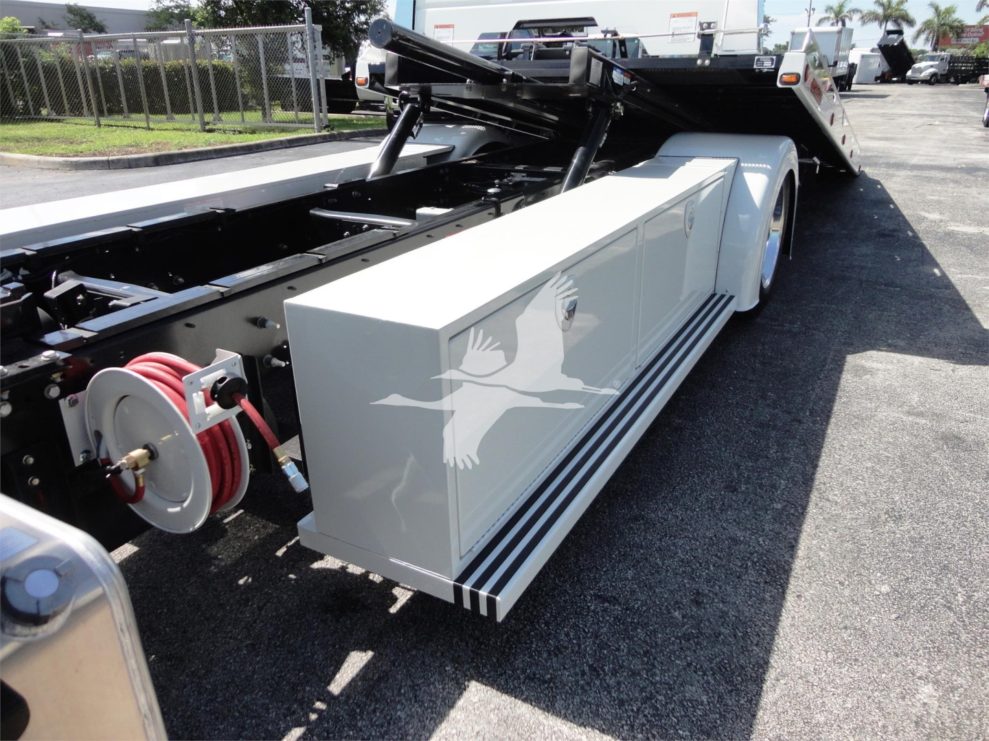 Truck body showing a white side storage box mounted on a rollback truck bed, with hoses and hydraulic reels visible and a raised bed in the background.