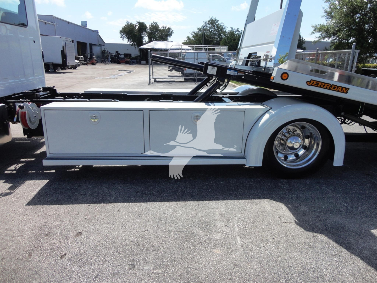 Side view of a white rollback tow truck with chrome wheel, storage compartments, and a folded riser bed with a Jerr-Dan branded rail at rear.