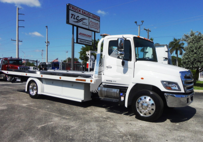 White 2022 Hino 268A rollback tow truck parked at a dealership lot with TLC Truck Sales Center sign in background.