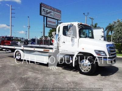 Flatbed tow truck in white with cab and car carrier, side view, dealership lot in background.