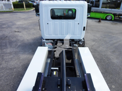 Rear view of a white 2022 Hino 268A roll-back truck. View shows the truck cab over with a black chassis and white side compartments extended; parking lot background with another green bus.