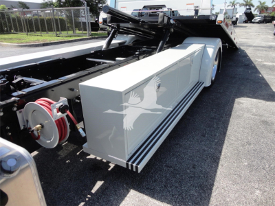 Truck body showing a white side storage box mounted on a rollback truck bed, with hoses and hydraulic reels visible and a raised bed in the background.