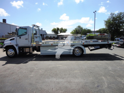 Side view of a 2022 Hino 268A rollback tow truck with a flatbed, white cab, and long silver bed in a dealership lot.