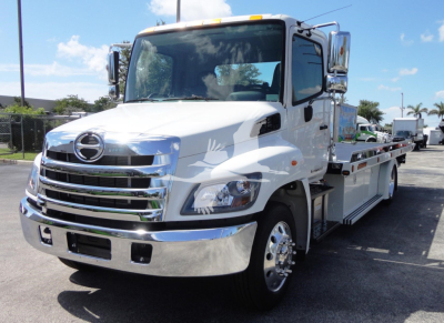 Tow truck featuring a white cab and chrome front, Hino badge, flatbed with wheel lift on a sunny day.