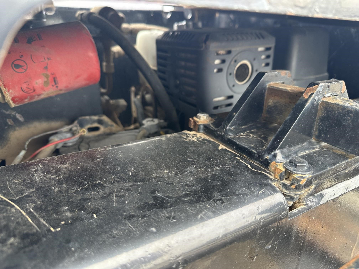Load King lowboy trailer engine bay area showing battery, red cap, and black components with some bolts and bracket; visible labels include no clear alphanumeric text readable.