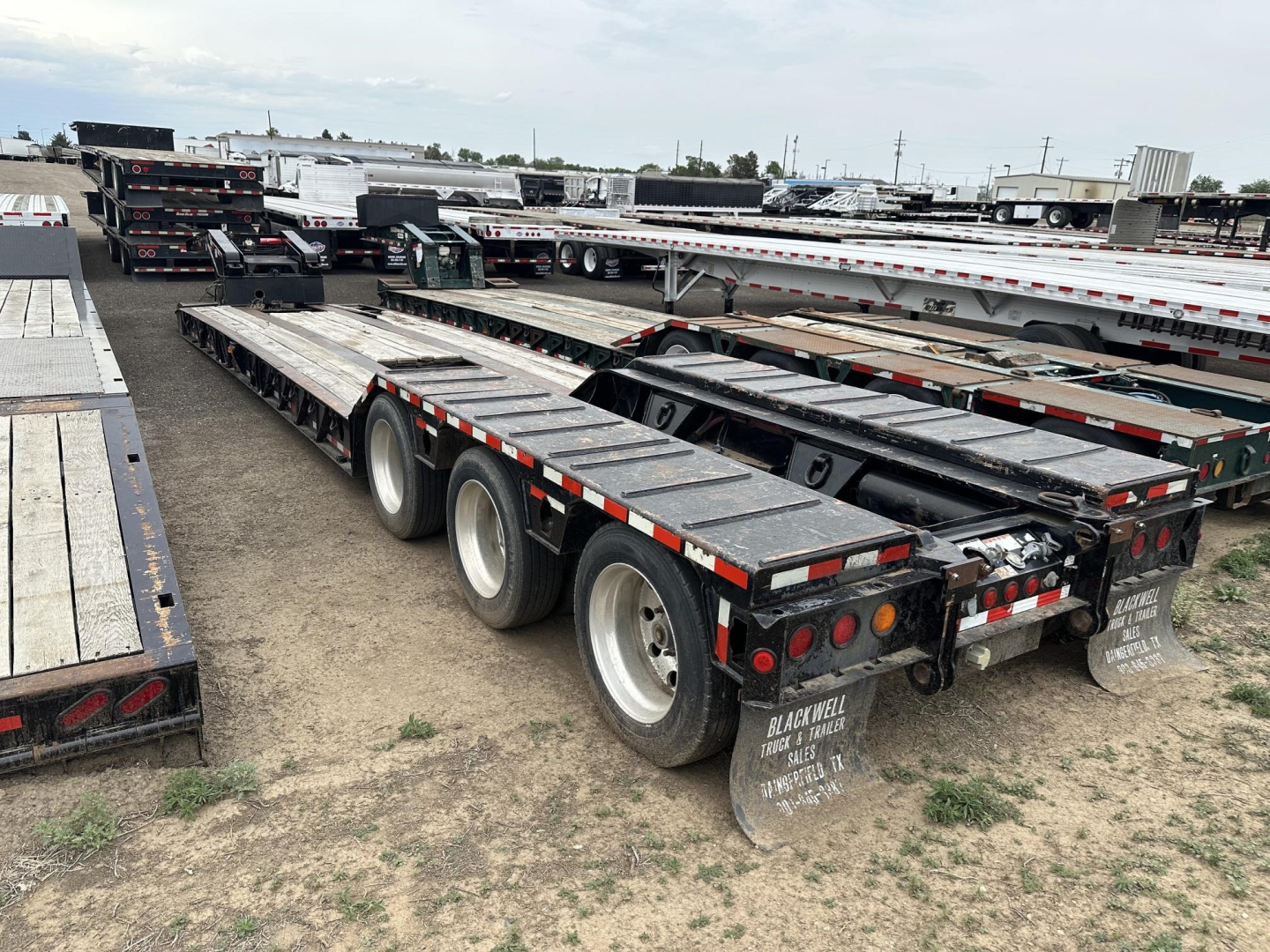 Trailer chassis with dual rear axles, Load King lowboy flatbed trailer in a used lot, with metal decking and rear mud flaps.