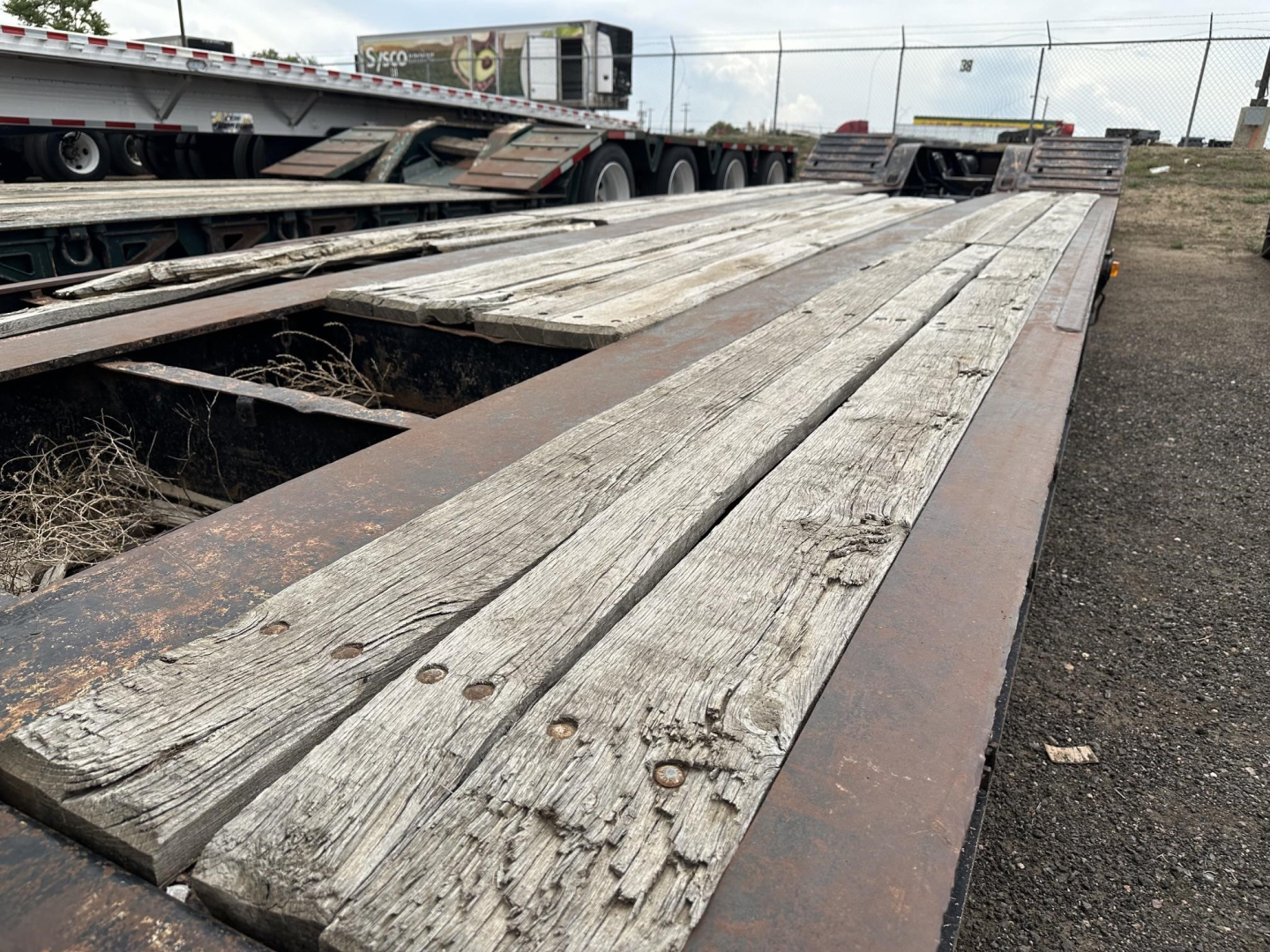 Trailer bed showing weathered wooden planks and steel frame of a lowboy trailer. Visible: long wooden deck boards with nails, rust on metal edges, gravel ground.