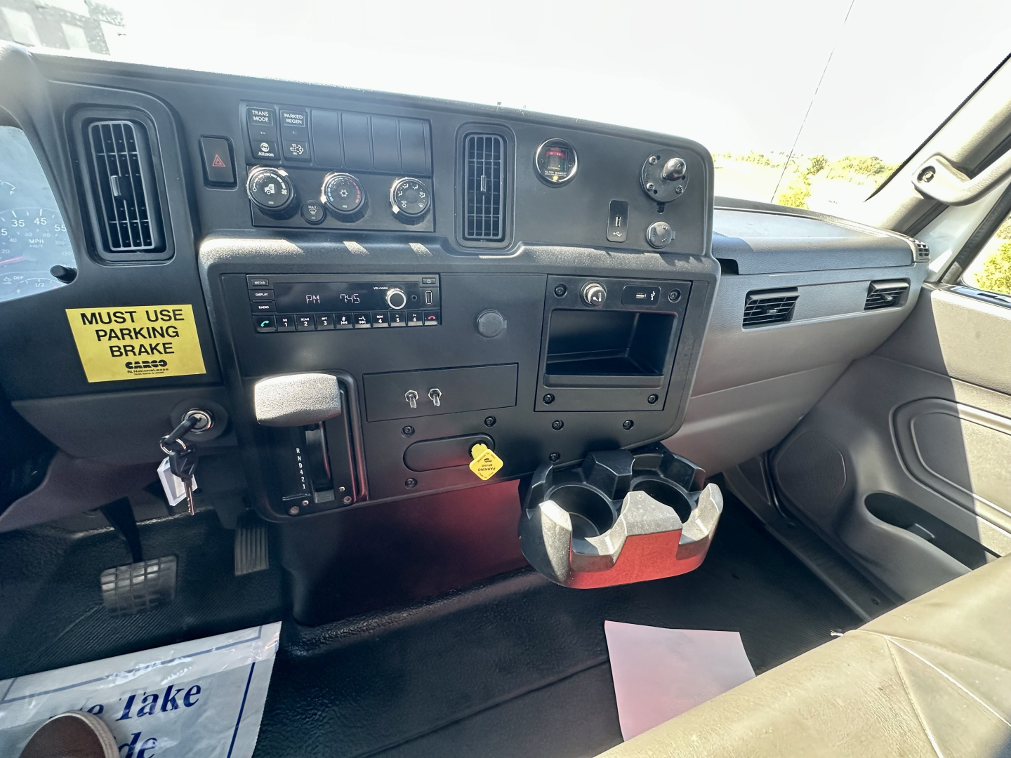 Truck cab dashboard of a 2023 International MV607 showing steering column, control switches, cup holders, ignition area, and yellow warning label near parking brake.
