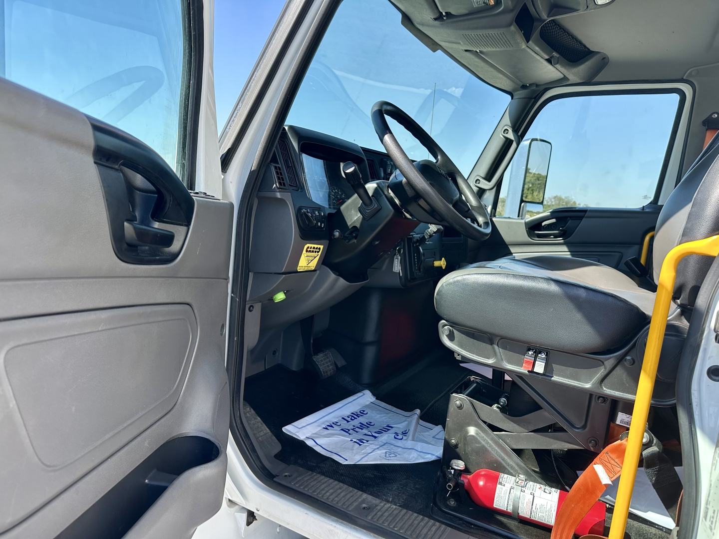 Truck cab interior of a 2023 International MV607 showing driver seat, steering wheel, dashboard, pedals, and door with a yellow handrail in view.