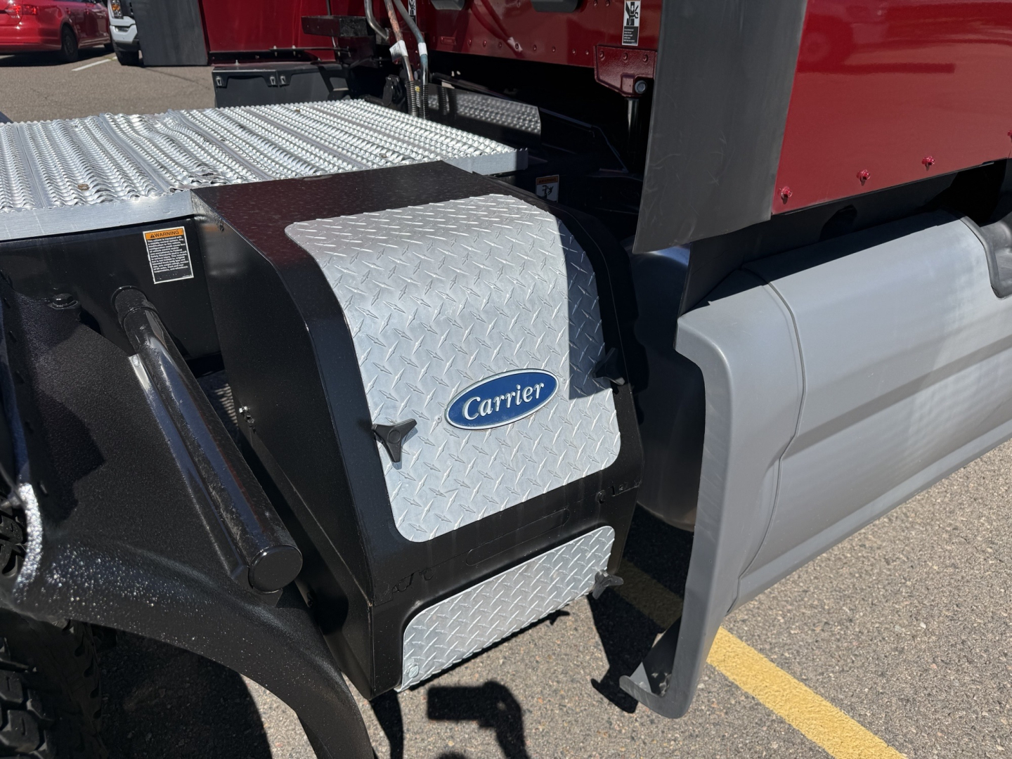 Truck step and Carrier fuel/air storage box mounted on the driver side of a 2021 Peterbilt 579 conventional sleeper truck; Diamond tread aluminum step; Carrier logo on a silver diamond-plate box; gray side fairing and hitch area visible.