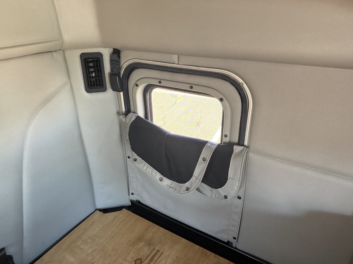Peterbilt 579 interior window well with fold-down window shade and light-gray upholstery; small vent on left; plywood floor visible.