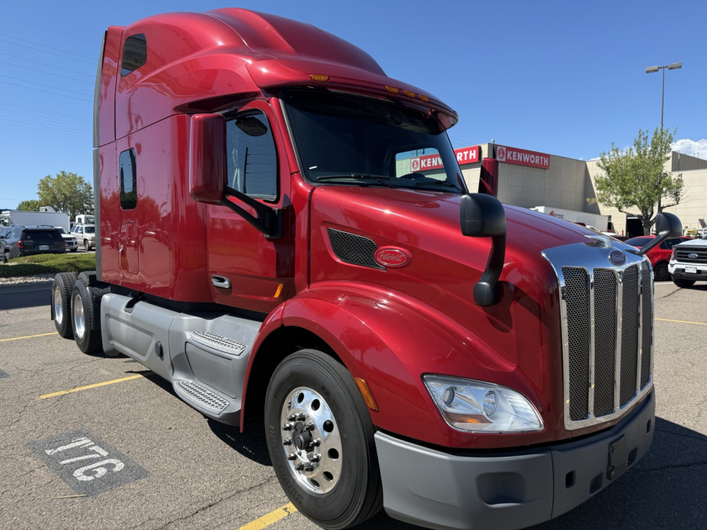 Peterbilt 579 conventional sleeper semi-truck in red, front-right view, chrome grille, dual rear axles, Kenworth dealership in background.