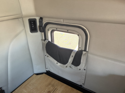 Peterbilt 579 interior window well with fold-down window shade and light-gray upholstery; small vent on left; plywood floor visible.