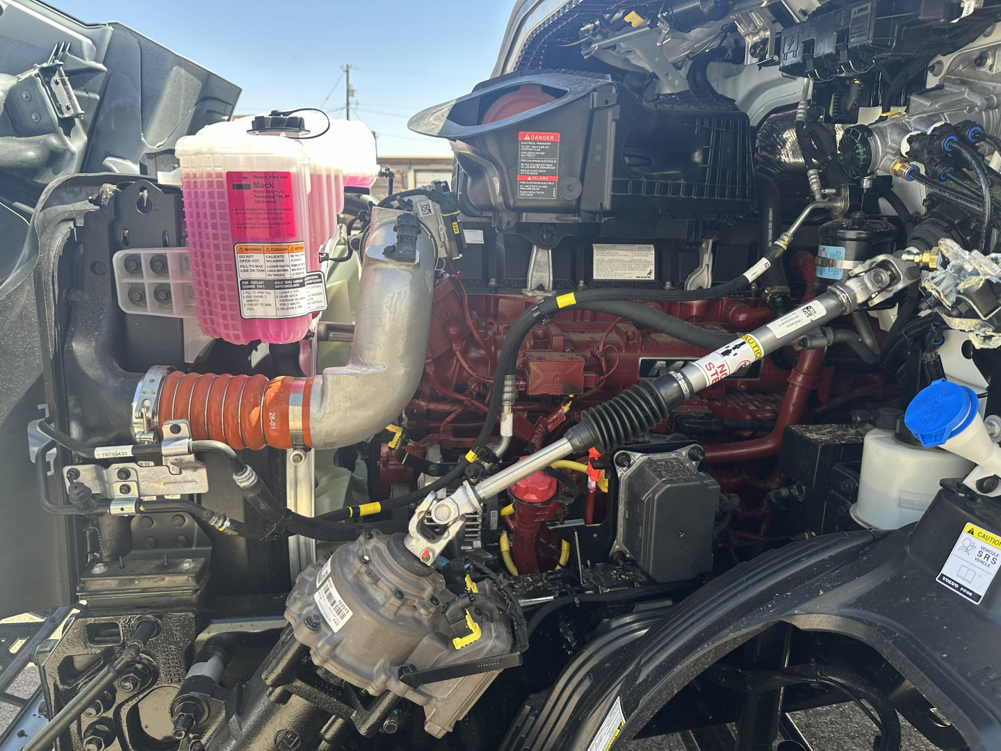 Truck engine bay showing pink coolant reservoir, red engine block, wiring, hoses, and blue cap in Mack AN64T engine area.