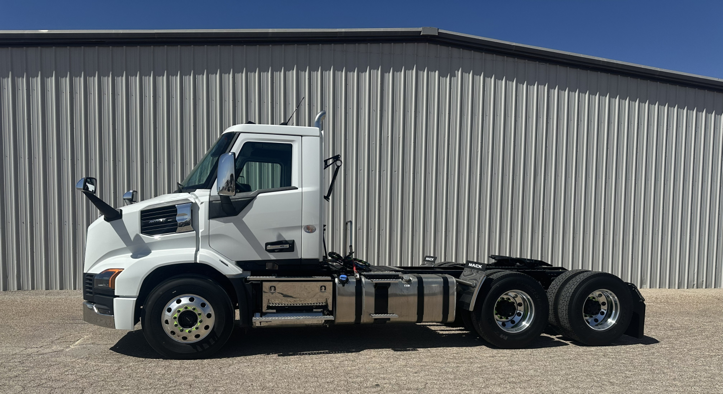 Truck side view showing cab and chassis of a new Mack AN64T conventional daycab with triple axle rear, white cab, black undercarriage.