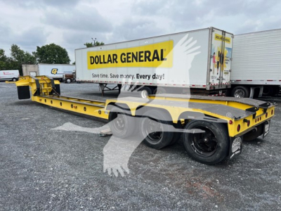 Limited view of a yellow 50 ton beam lowboy trailer with dual rear axles and a Dollar General logo on a nearby trailer.