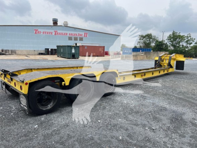 Rogers 50 Ton beam trailer in yellow, long flatbed with rear axles and gooseneck, side rails, trailer hitch area, industrial yard with building in background.
