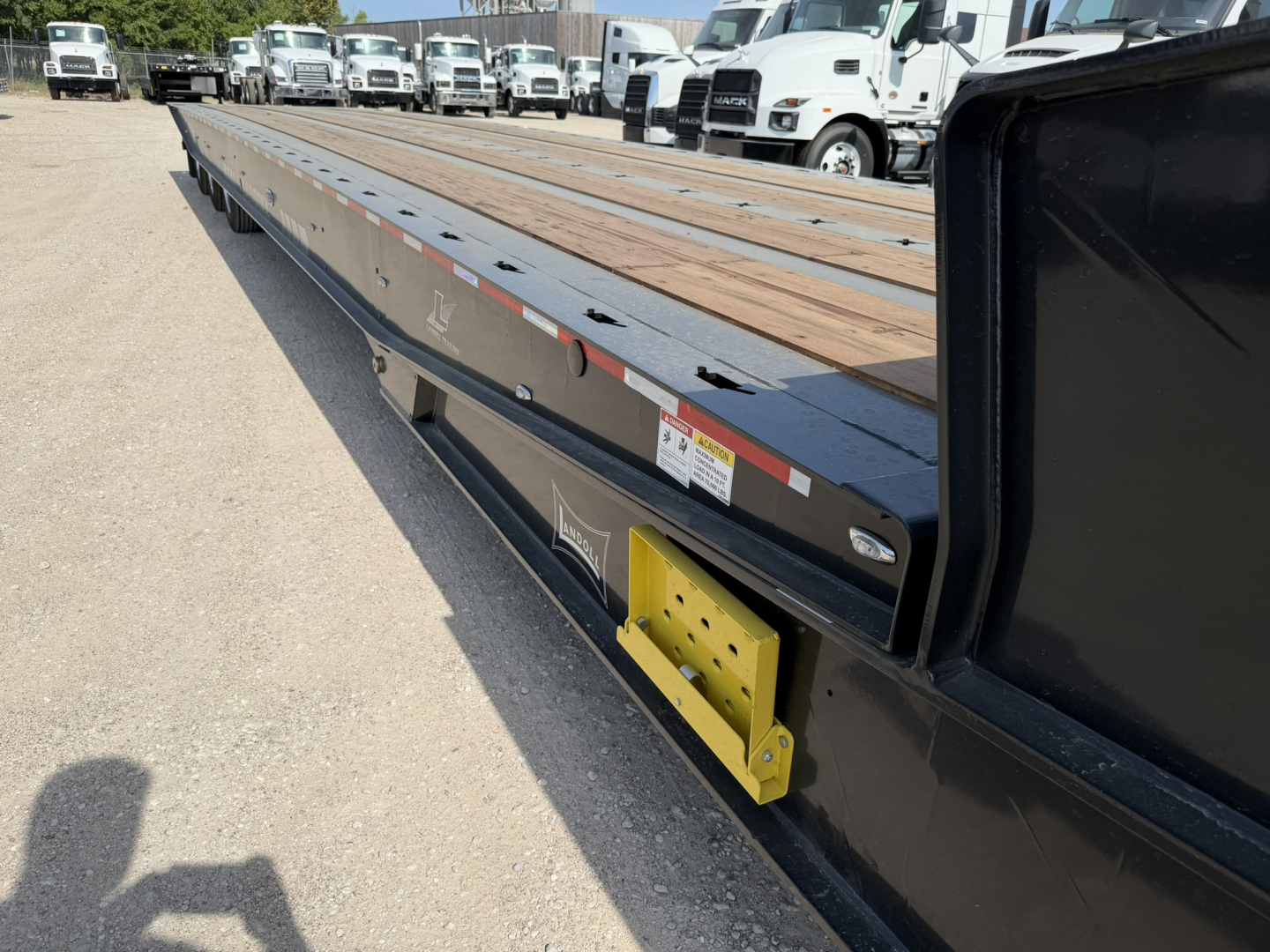 Long lowboy trailer with wooden deck and black sides; side of trailer shows yellow bracket and various stickers; several trucks in background in a lot