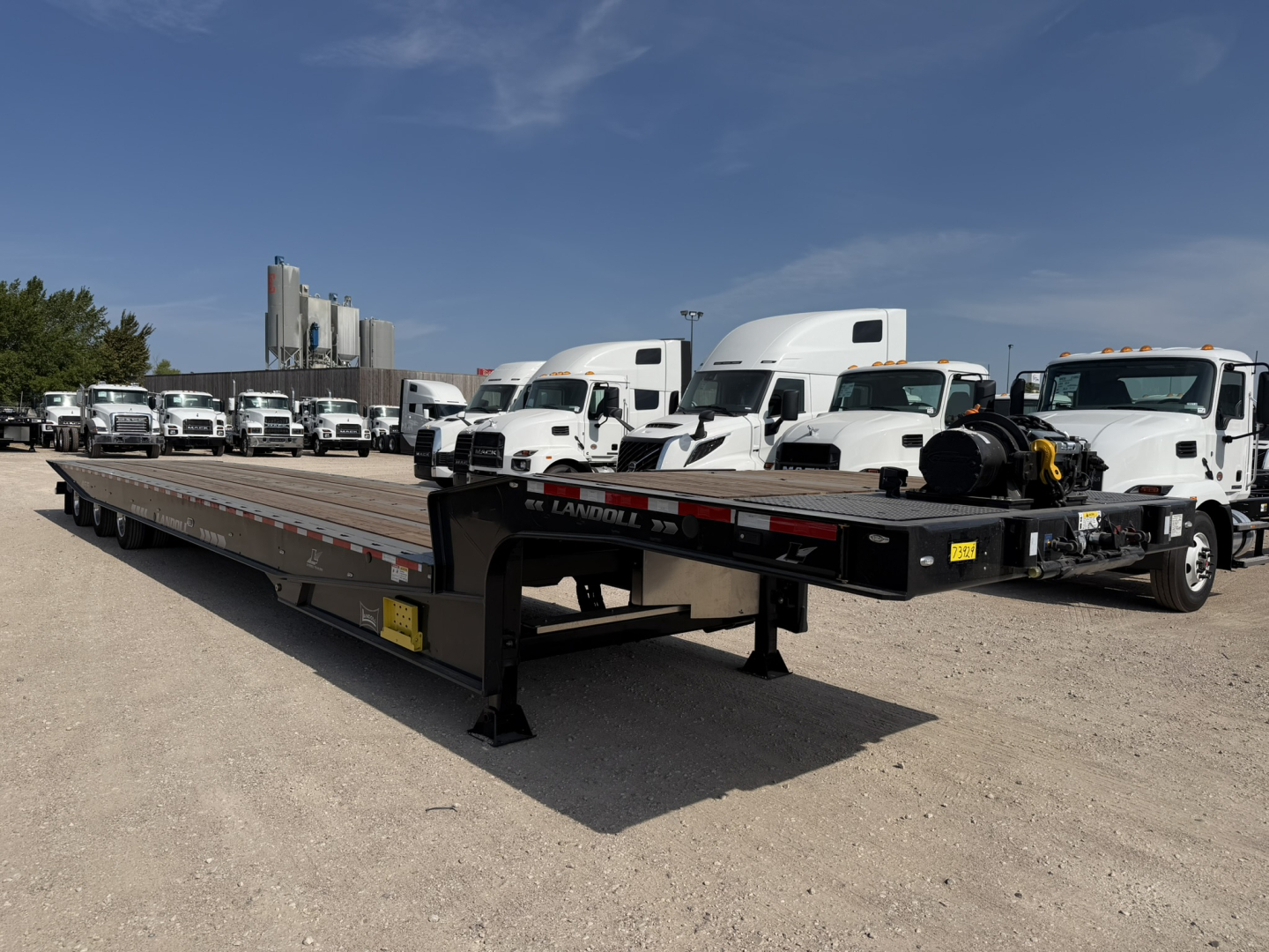 Lowboy trailer with extended flatbed and gooseneck showing Landoll logo, 55-ton capacity, amid a row of white trucks in a dealership lot.