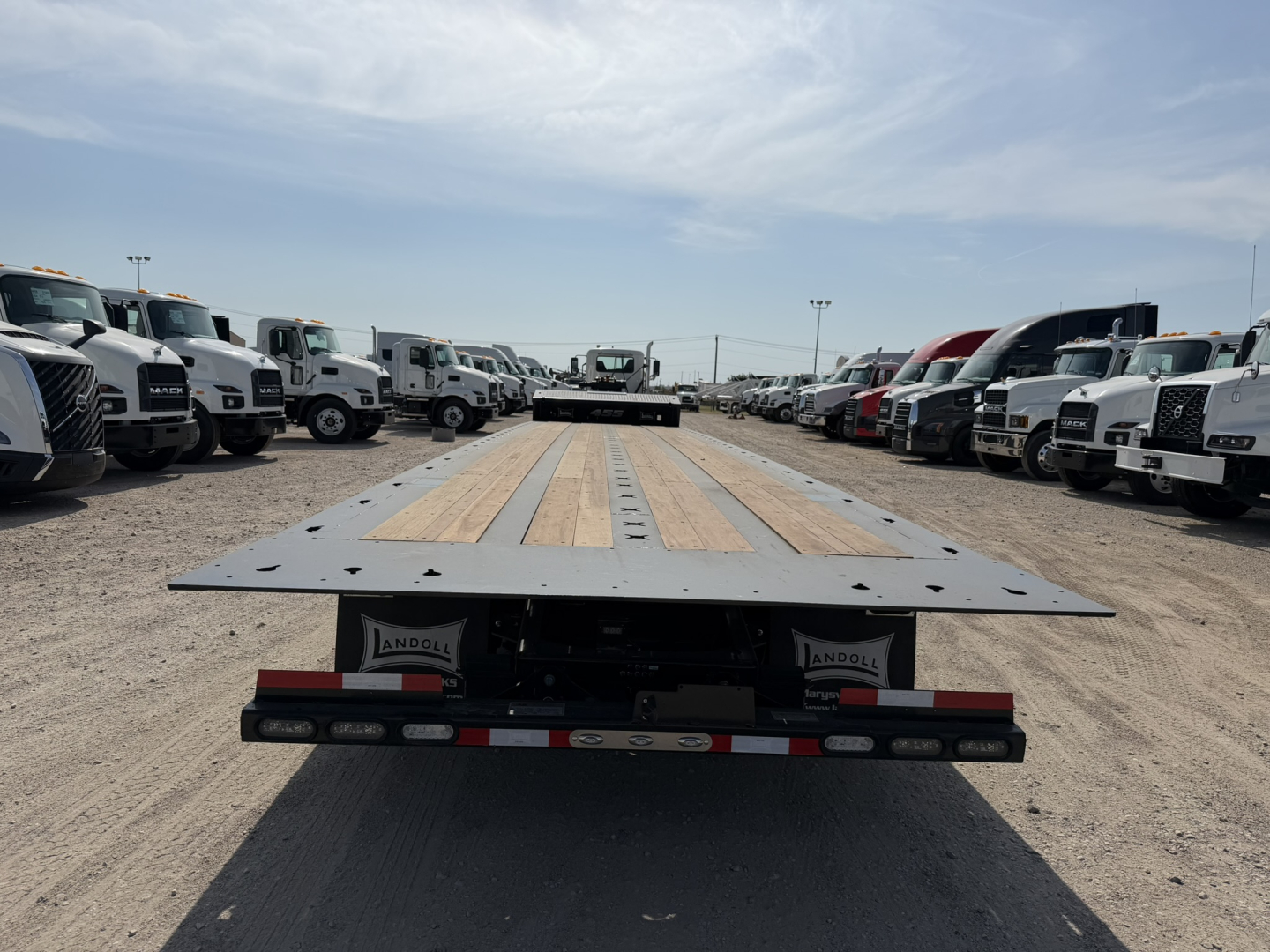 Flatbed trailer deck of a Landoll 455B-53 55-ton traveling axle, viewed from the rear, with wood deck and metal frame, line of trucks on both sides.