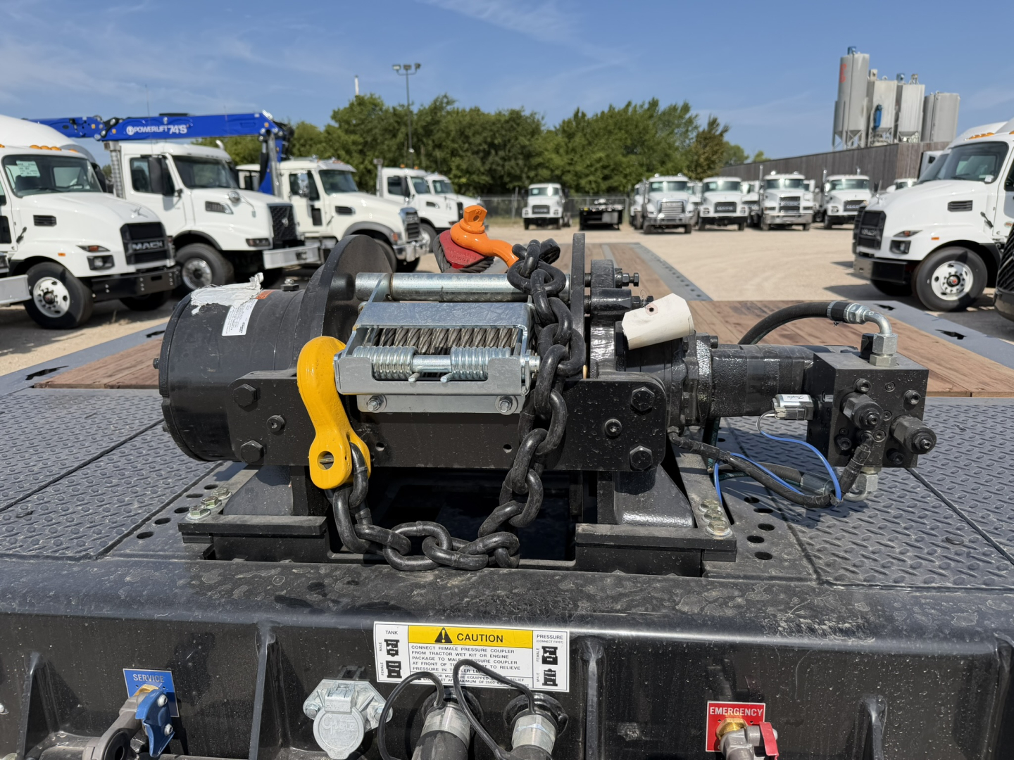 Tow trailer winch and hydraulic system mounted on a flatbed, with chain and yellow hook, warning label, and control levers visible.