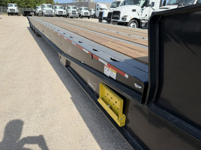 Long lowboy trailer with wooden deck and black sides; side of trailer shows yellow bracket and various stickers; several trucks in background in a lot