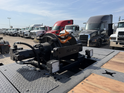 Traveling axle 55 ton Landoll trailer hitch mechanism with hydraulic winch and metal diamond-plate deck, various trucks in background.