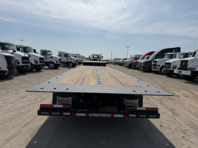 Flatbed trailer deck of a Landoll 455B-53 55-ton traveling axle, viewed from the rear, with wood deck and metal frame, line of trucks on both sides.