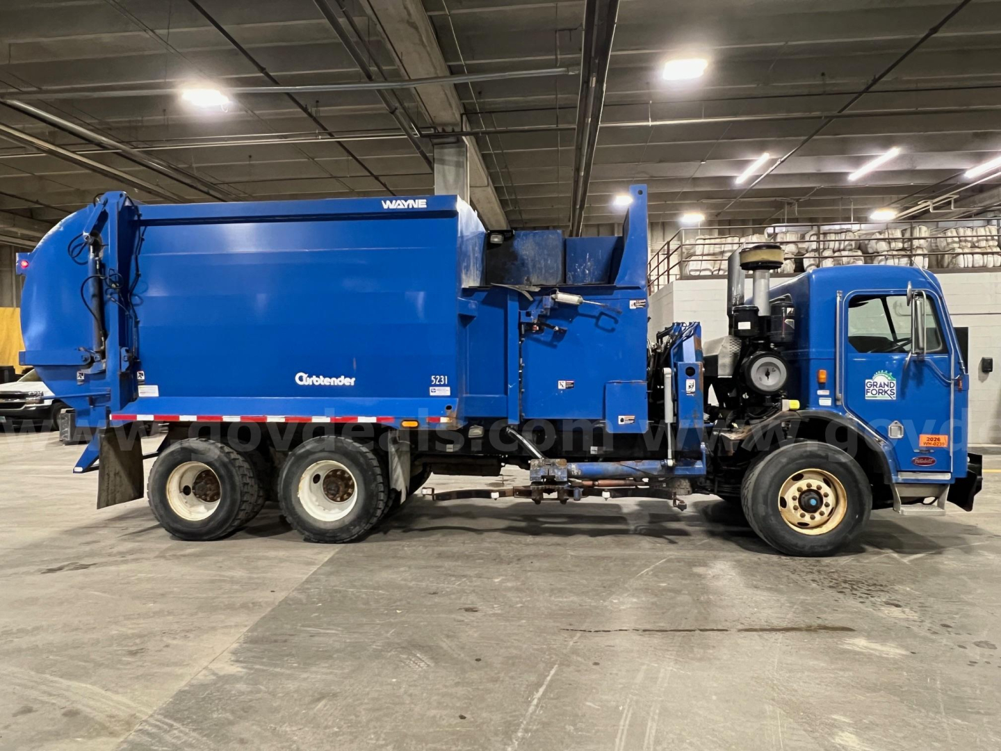 Peterbilt 320 garbage truck in an indoor facility, side view showing blue cab and rear-compactor body with lettering 'Vogone' and 'Grapen' logos, VIN plate area not legible.