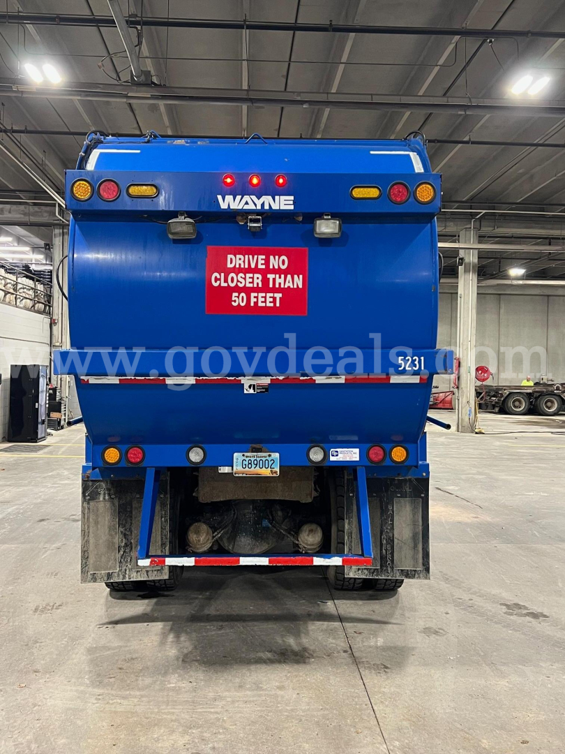 Peterbilt 320 rear view garbage truck in blue with Wayne body, red warning sign, license plate, and stock number plate in an indoor facility.