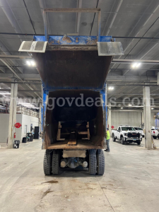 Dump truck rear view showing open bed with blue chassis, raised tailgate, and dual rear wheels; visible rust and support structure under the bed.