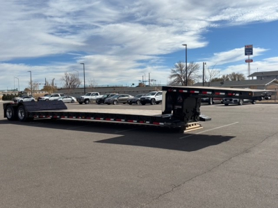 Flatbed trailer with dual drop legs and swing-out ramps in a dealership lot; long black trailer with red/white reflective tape along side, front hitch area visible