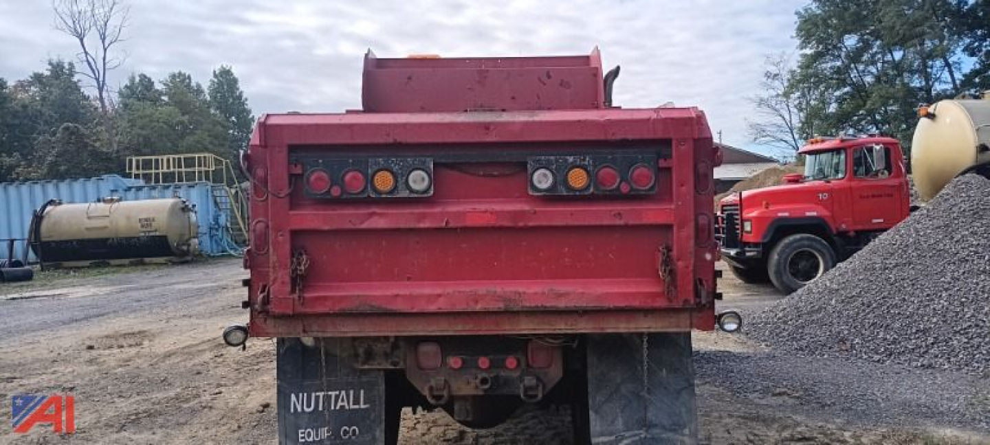 Rear view of a red dump truck bed with taillights and hitch, vintage Ford chassis, in a gravel yard with other trucks and a large yellow tank in the background.