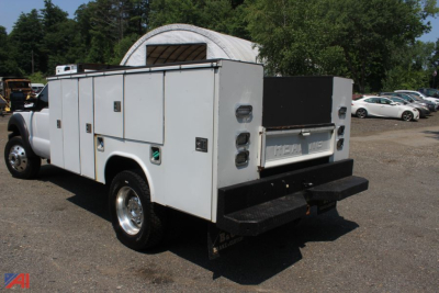 Ford F-550 utility service body truck seen from rear-left, white service compartments along sides, black rear bumper, dual rear lights, cabin visible, gravel lot with trees and other vehicles.
