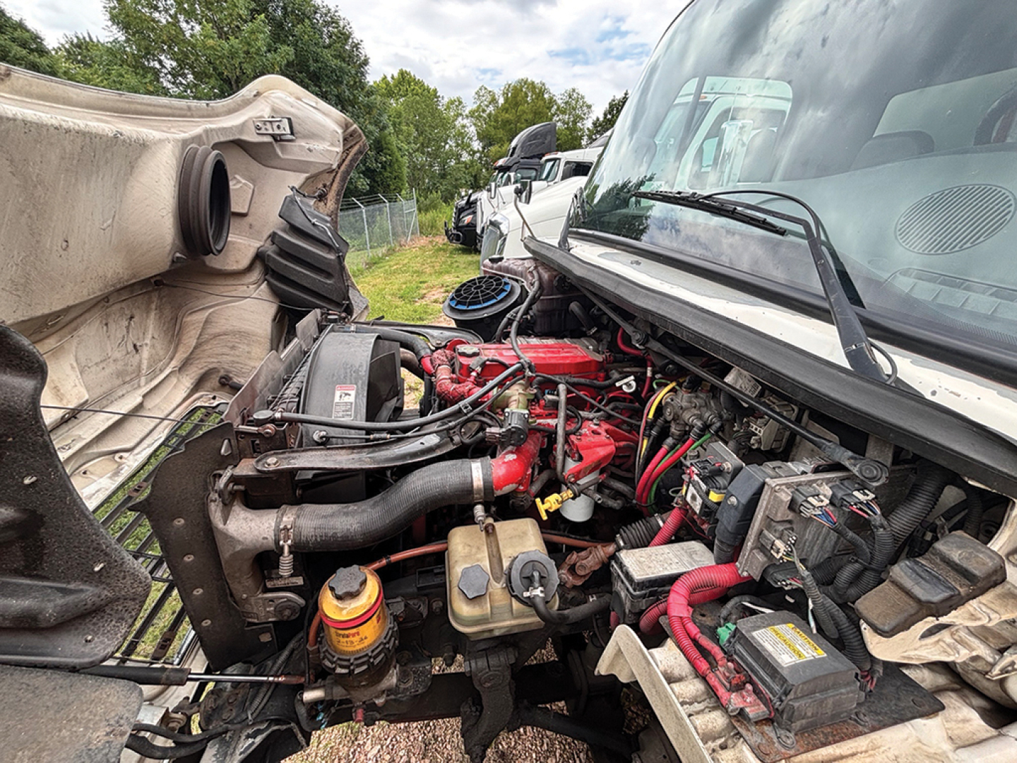 Truck engine bay view of a 2012 Freightliner M2 106 Business Class showing red engine components, coolant reservoir, battery, hoses, and surrounding engine bay.