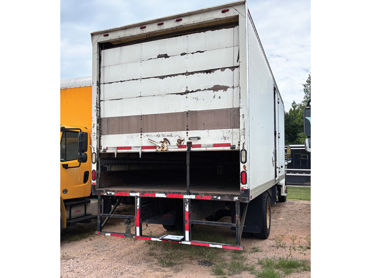 Truck rear view of a Freightliner M2 106 box trailer with raised roll-up door and metal steps, white with weathered panels.