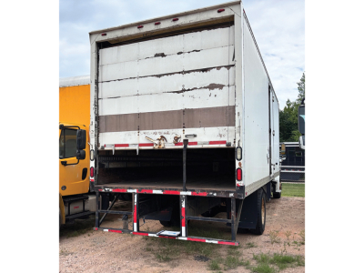 Truck rear view of a Freightliner M2 106 box trailer with raised roll-up door and metal steps, white with weathered panels.