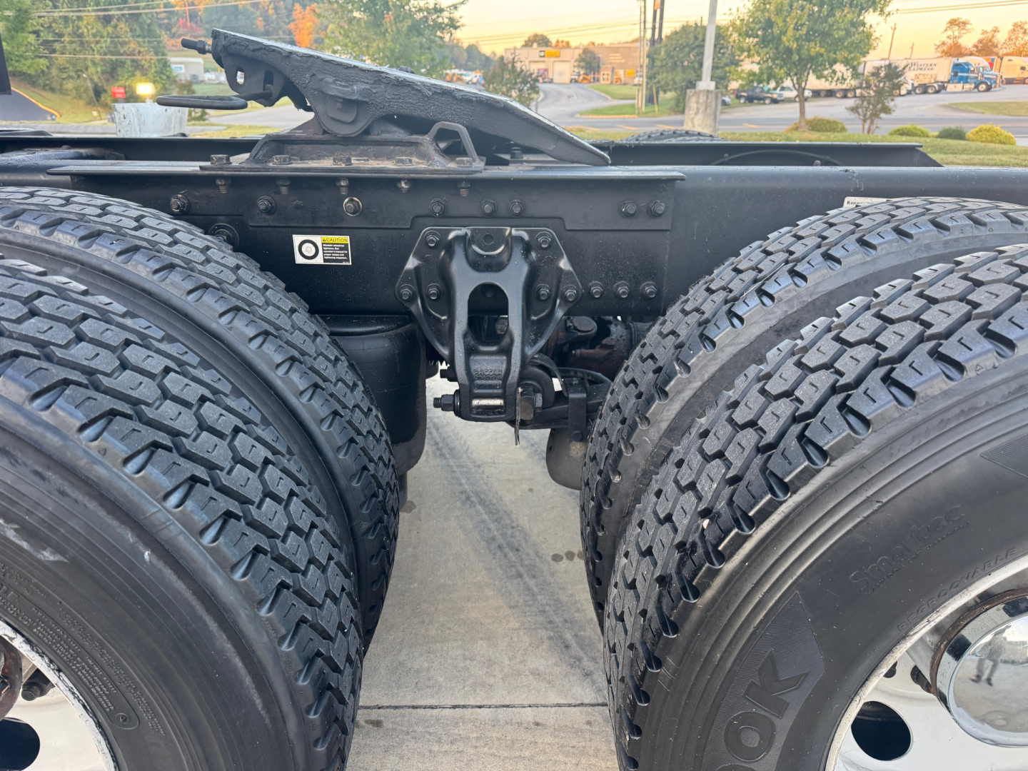 Kenworth T680 chassis view showing dual rear axles and fifth-wheel mounting area with various labels. Visible text includes warning label on left side and structural components.