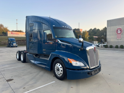 Kenworth T680 sleeper cab truck in blue showing front grille, headlights, side mirrors, and dual rear axles in a dealership lot.