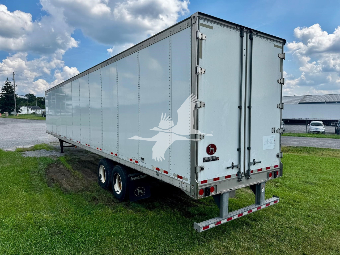 Flatbed trailer with enclosed silver box, triple-axle wheels, rear barn doors, Great Dane plate van design, on grassy area under blue sky.