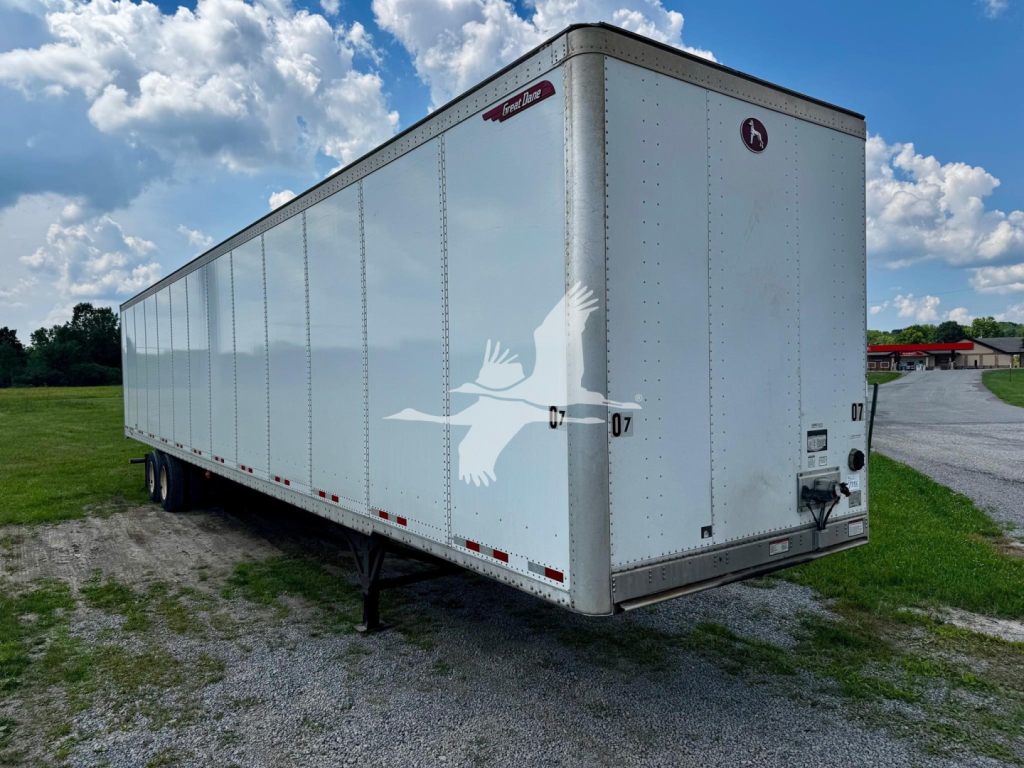 Trailer side and rear view showing a white Great Dane plate trailer with a rear doors, wheel area, and a small emblem near the top corner.