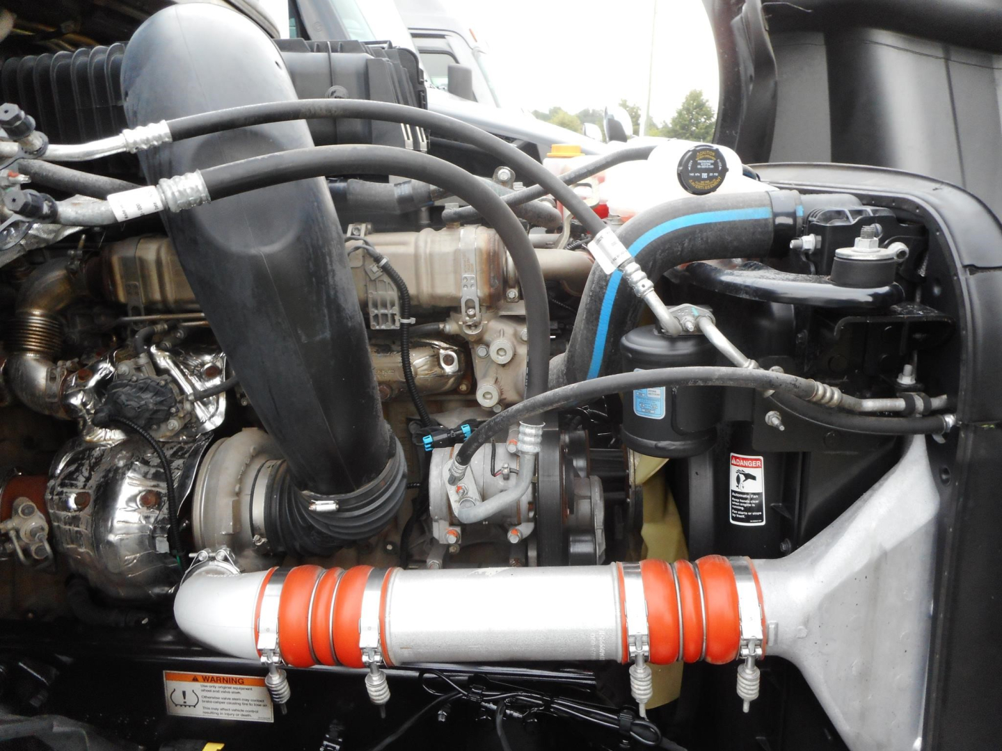Truck engine bay of a 2022 Freightliner Cascadia 126 showing intake hose, turbo, coolant reservoir with blue hose, orange silicone couplers on a silver pipe, and various hoses and components.