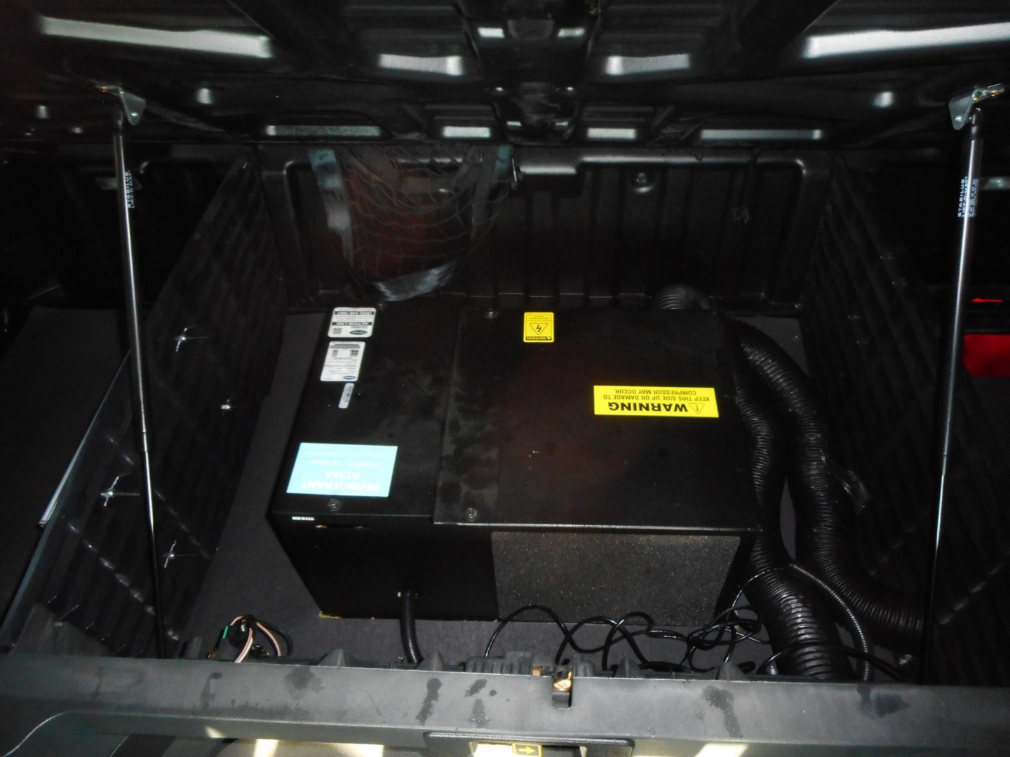 Trunk or engine bay of a freightliner Cascadia showing a black rectangular component with yellow warning label and blue sticker, wiring harnesses nearby.