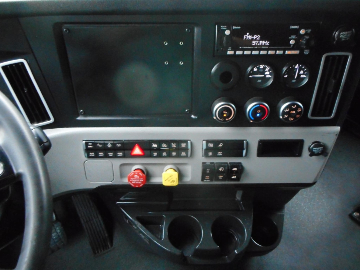 Freightliner Cascadia dash panel with radio display, gauges, climate controls, hazard button, and switches; two cup holders and storage below.