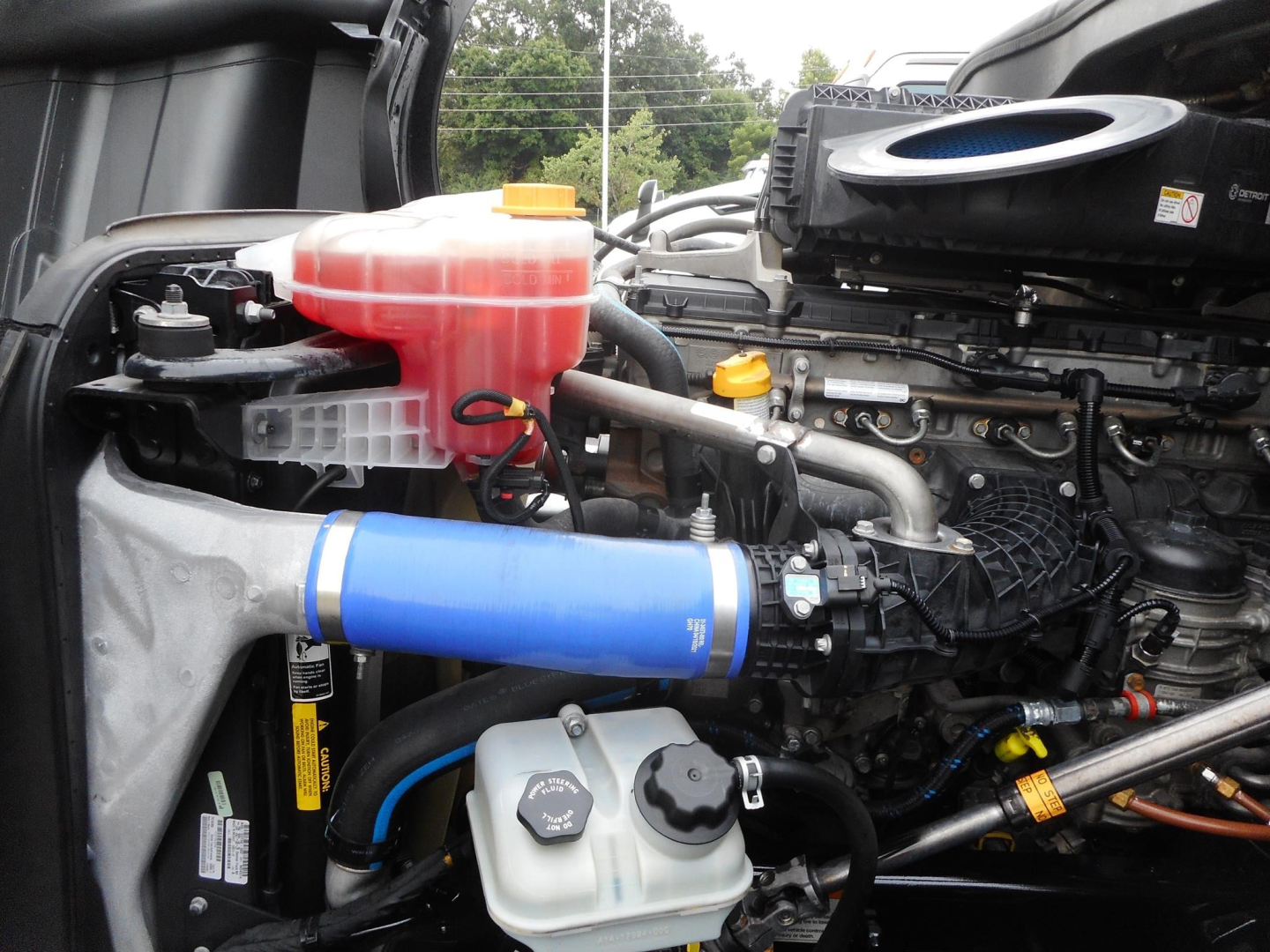 Engine bay of a Freightliner Cascadia showing blue silicone intercooler pipe, pink coolant reservoir, white coolant spill tank, various hoses, and engine components.