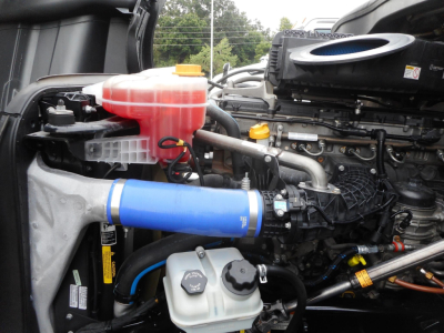 Engine bay of a Freightliner Cascadia showing blue silicone intercooler pipe, pink coolant reservoir, white coolant spill tank, various hoses, and engine components.